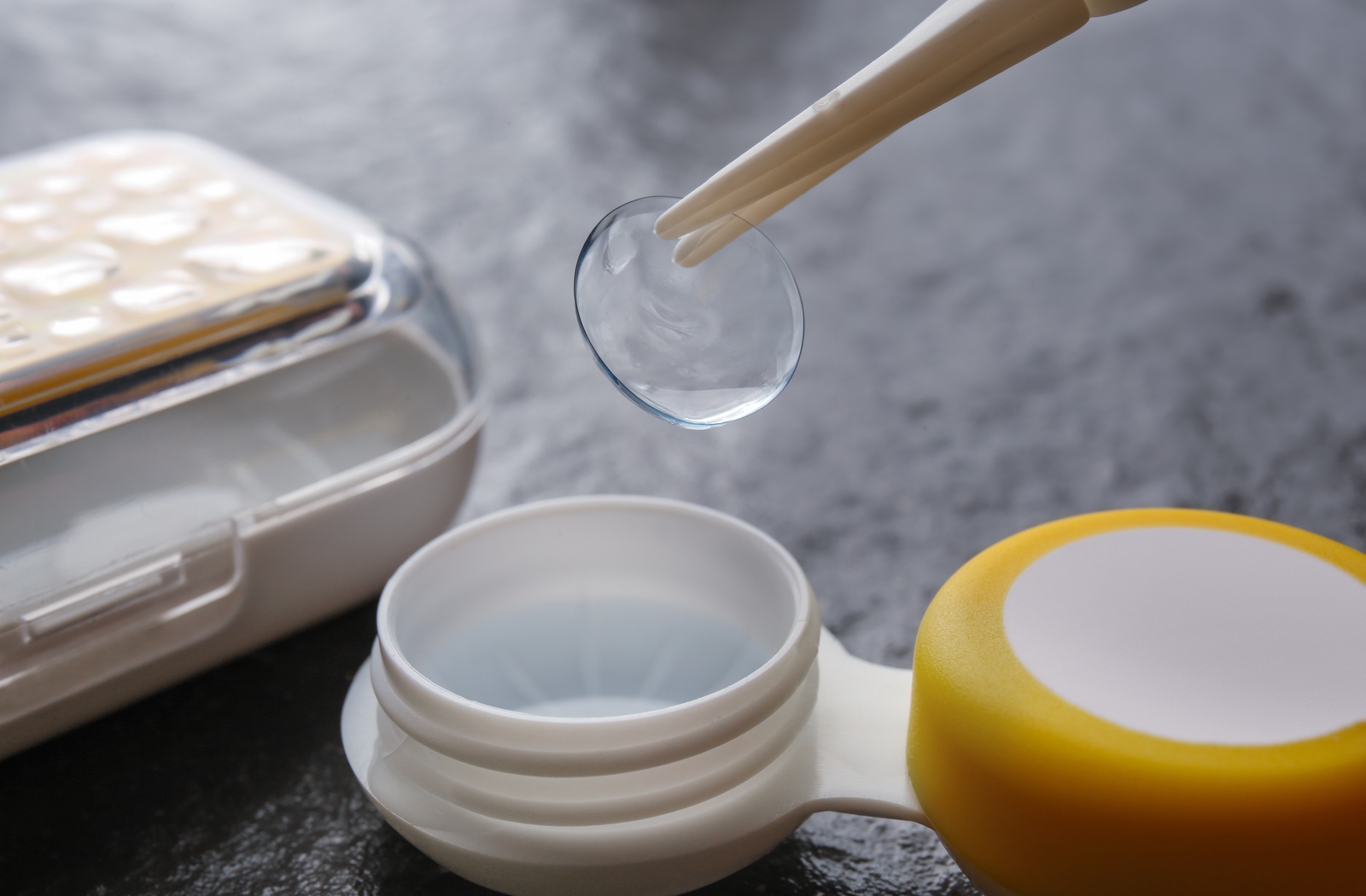 Someones using a pair of tweezers to take out a contact lens from it's plastic container