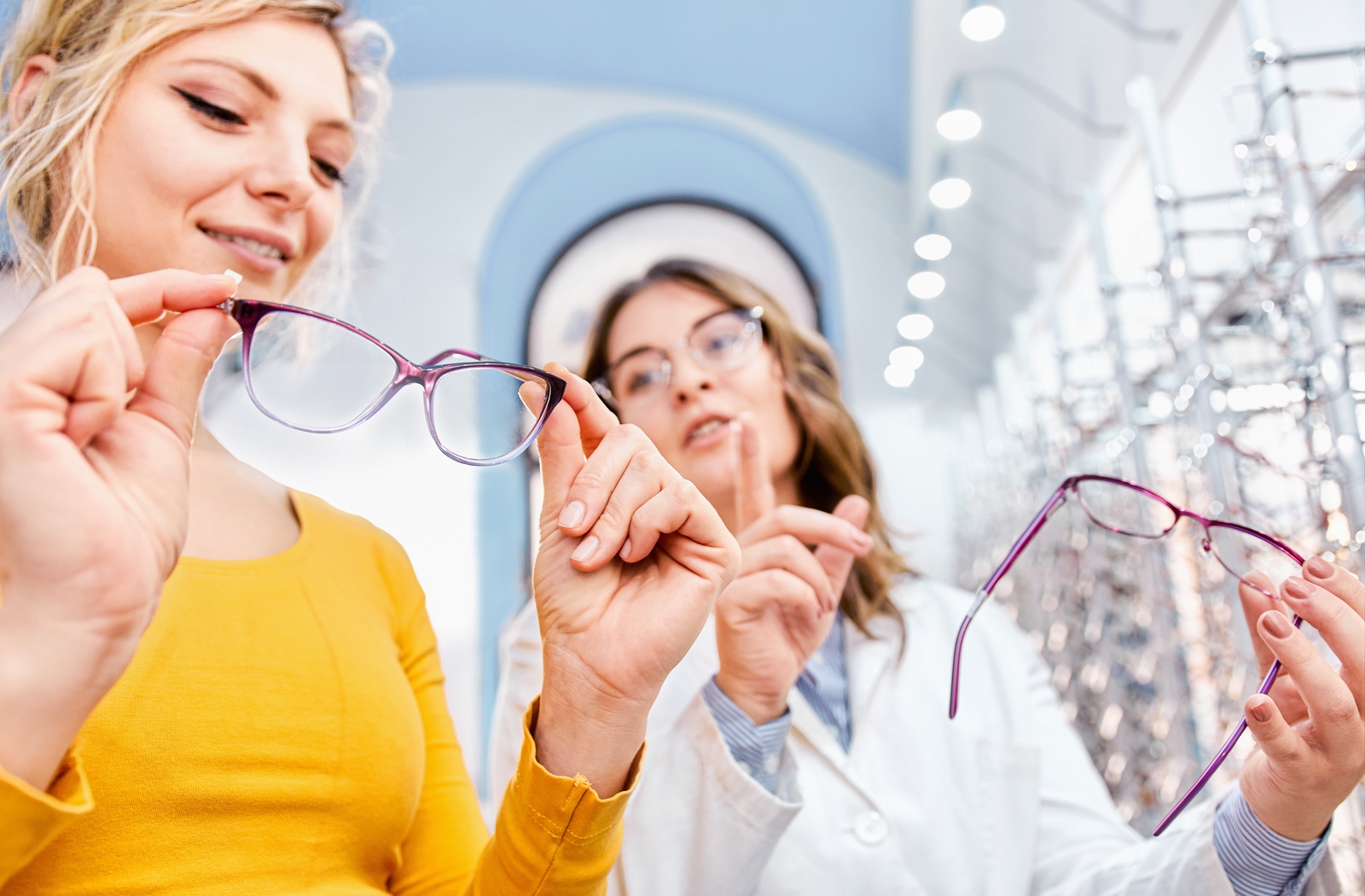 A female optician helping a woman with a bright yellow shirt pick out a new pair of glasses, while they both hold different pairs for her to try on