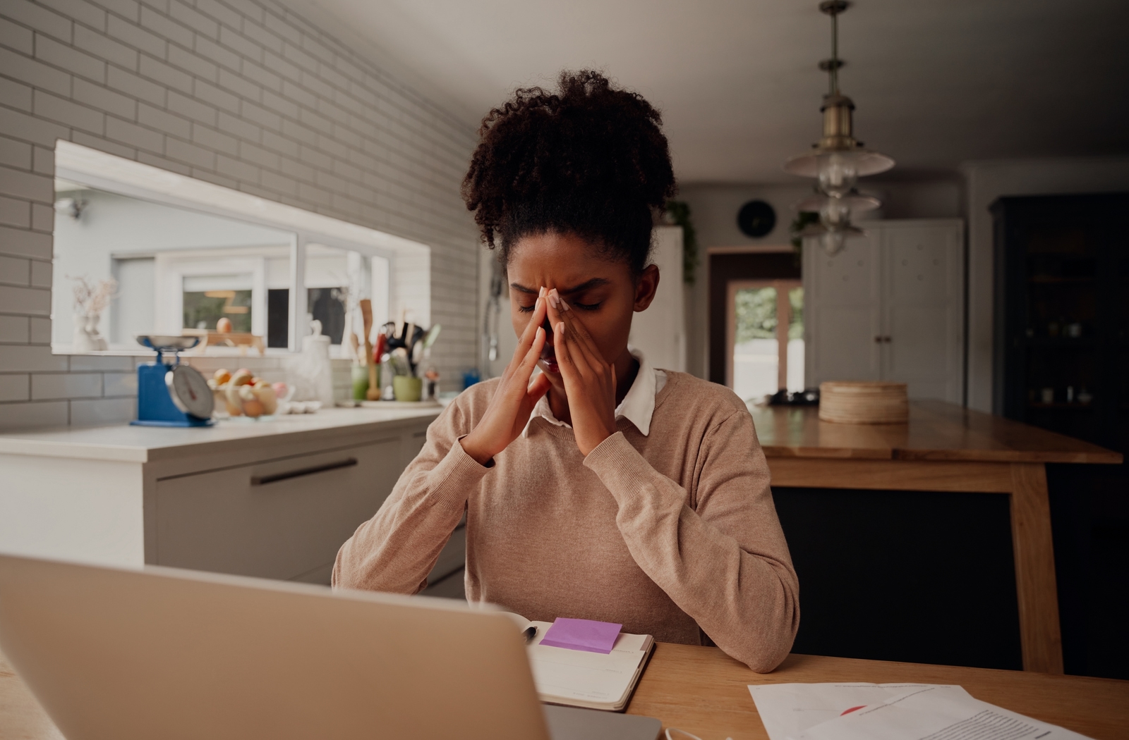 A woman sitting at a desk beside her kitchen, using her laptop, touching her eyes with both hands in discomfort due to her dry eye symptoms