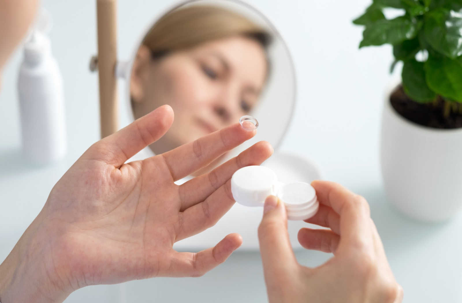 A mirror showing a reflection of a woman looking down at a contact lens on her finger and a contact lens case in her hand.