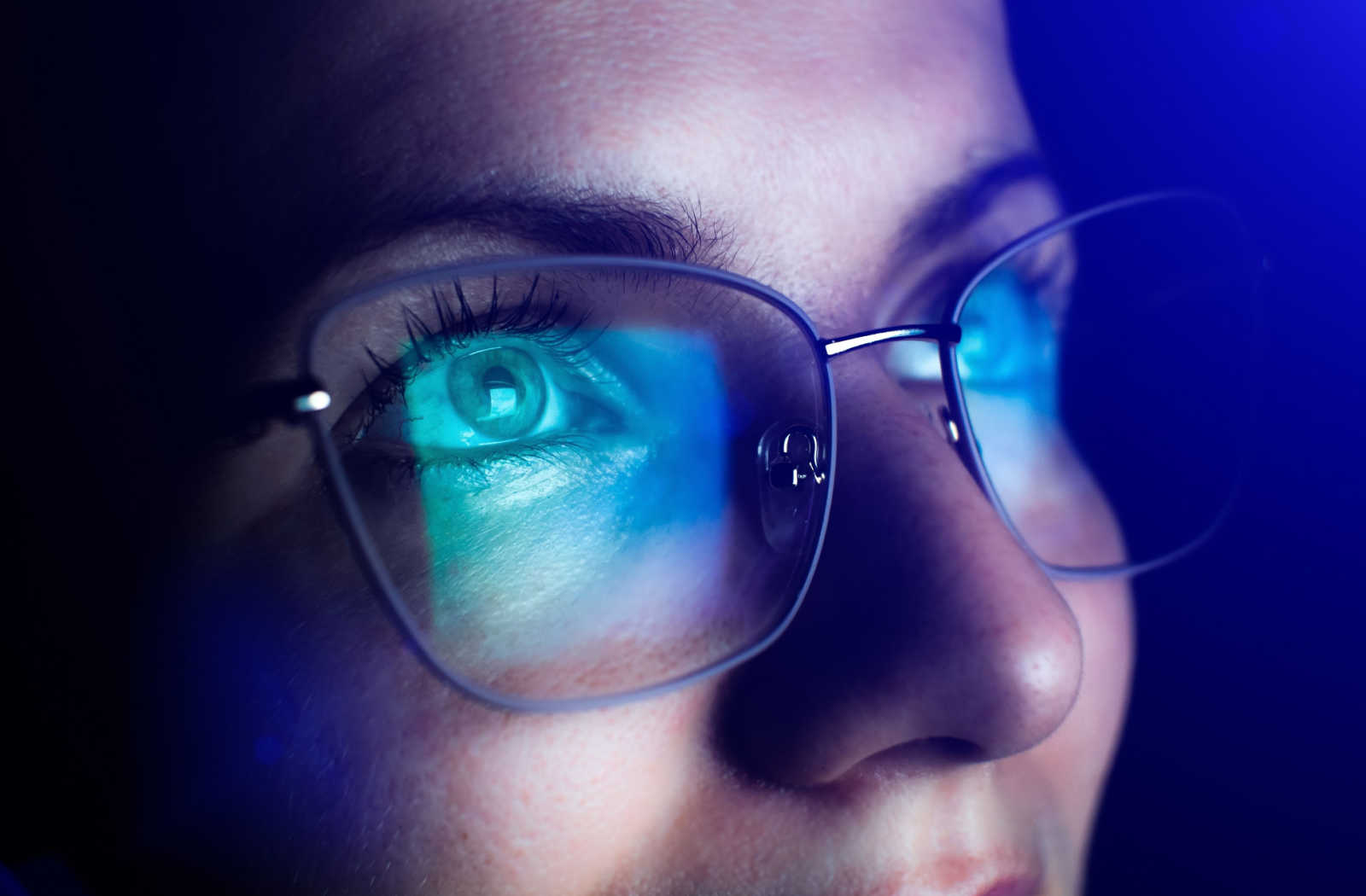 A close up of a woman wearing blue light glasses with a reflection of a digital screen on the lenses