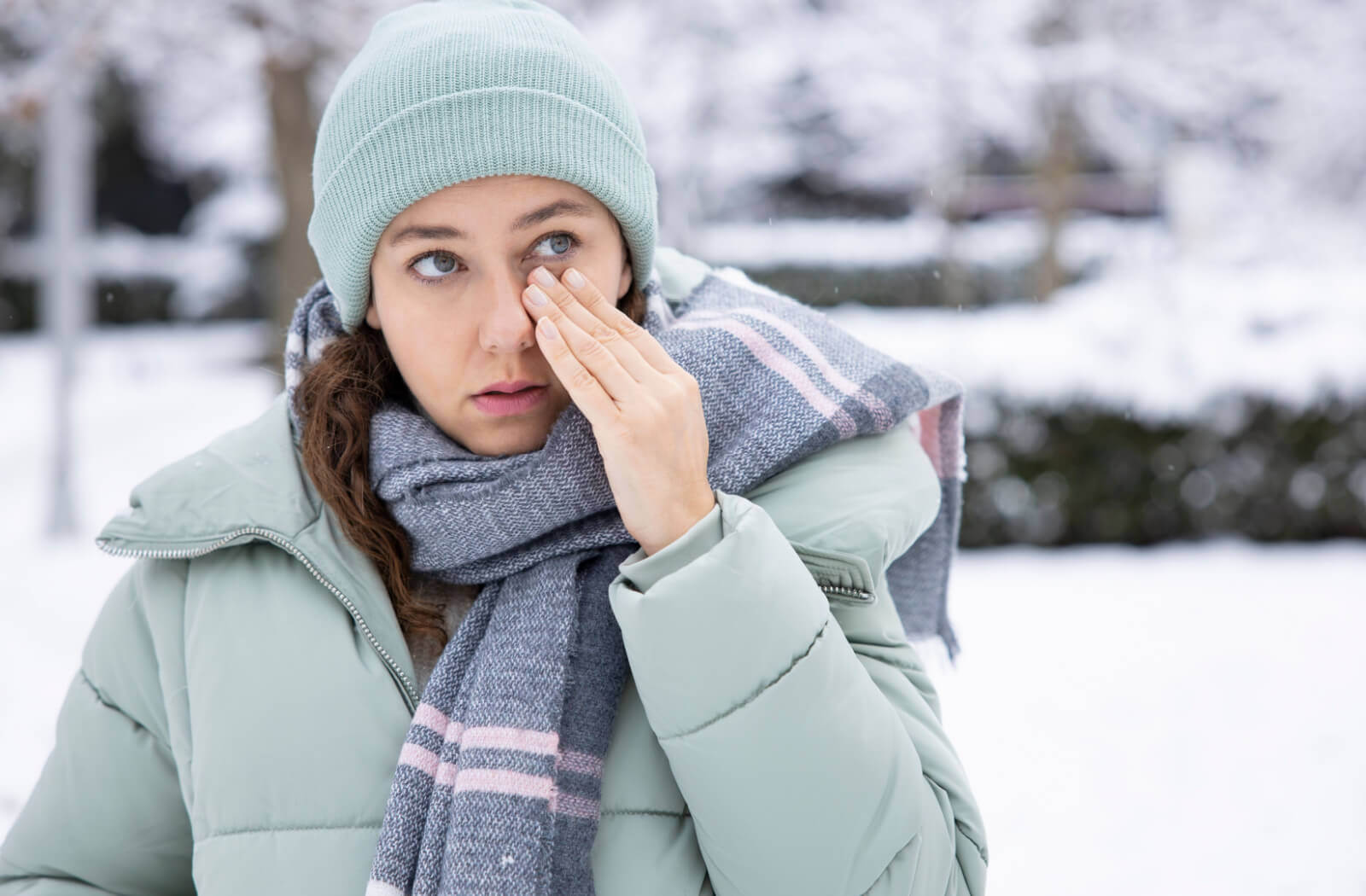 A woman in winter clothes outdoors is touching her left itchy eye.