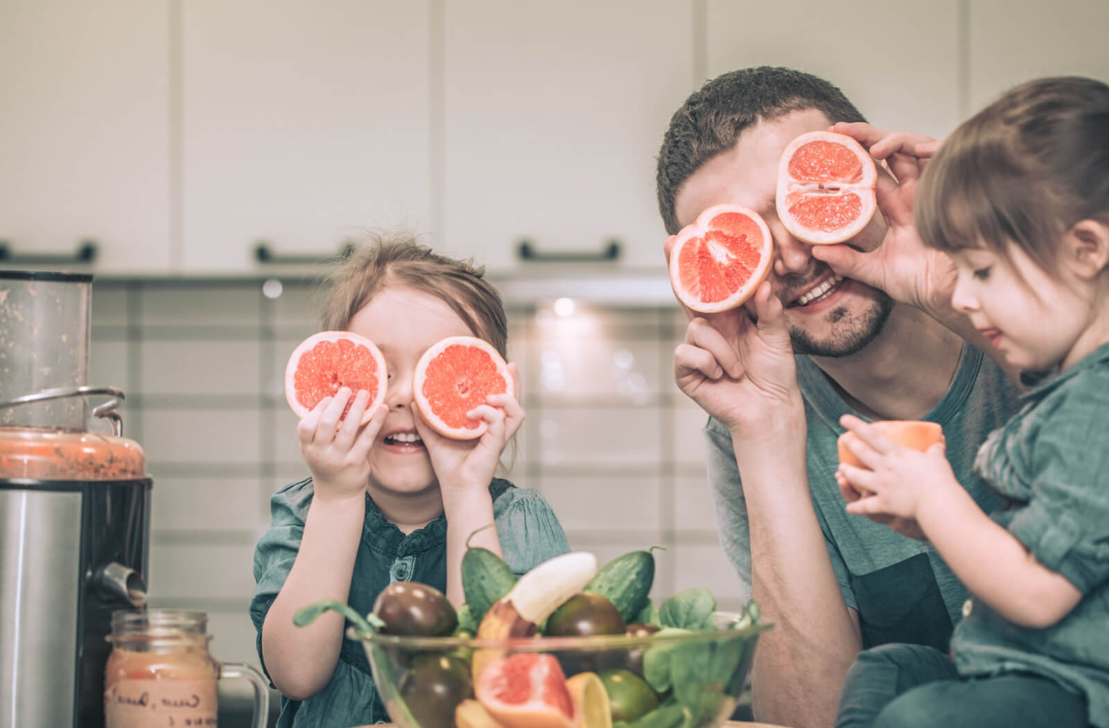 A father with his two lovely daughters prepares fresh juice from citrus fruits and vegetable in the kitchen .