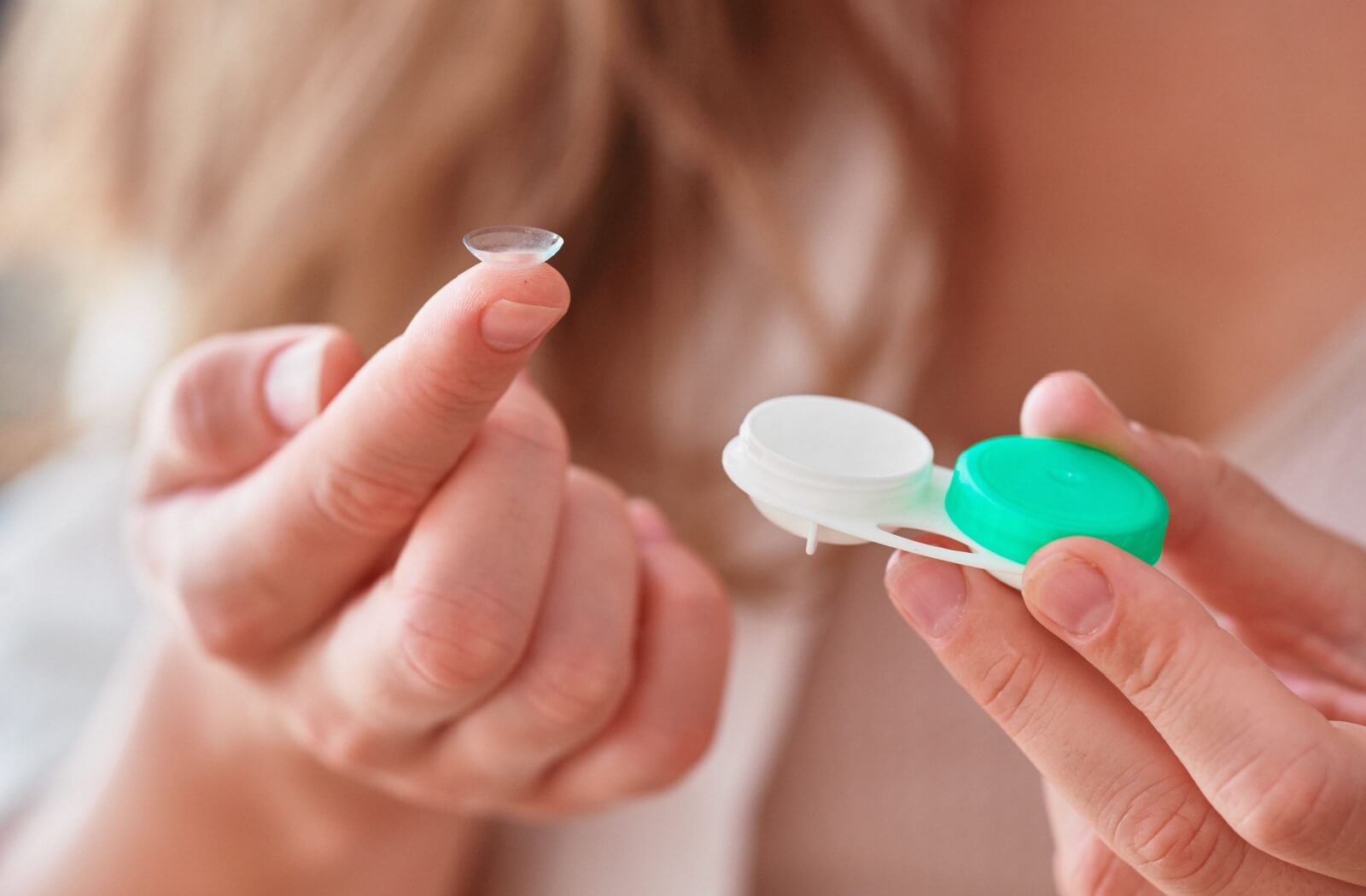 Close-up of a hand holding a contact lens case in one hand and a contact lens on a fingertip in the other.